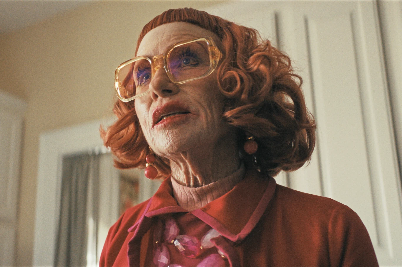 Amy Madigan w filmie "Zniknięcia" Starsza kobieta z rudymi, kręconymi włosami ubrana w czerwony sweter, różowy naszyjnik i duże okulary z żółtymi oprawkami, patrzy w górę w jasnym pomieszczeniu; w tle białe drzwi i fragment ściany.