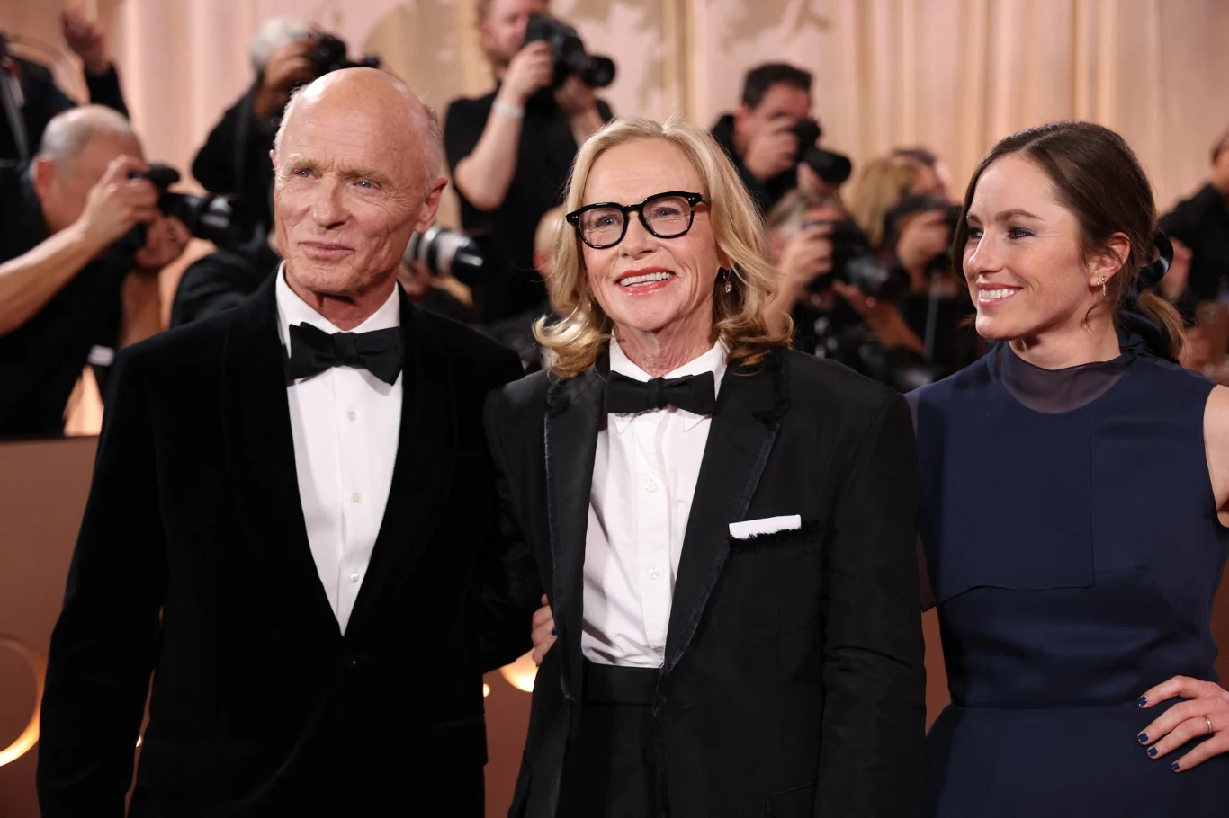 Ed Harris, Amy Madigan i ich córka - Lily Trzy uśmiechnięte osoby w eleganckich strojach stojące razem na tle jasnej dekoracji, w tle fotografowie wykonują zdjęcia.