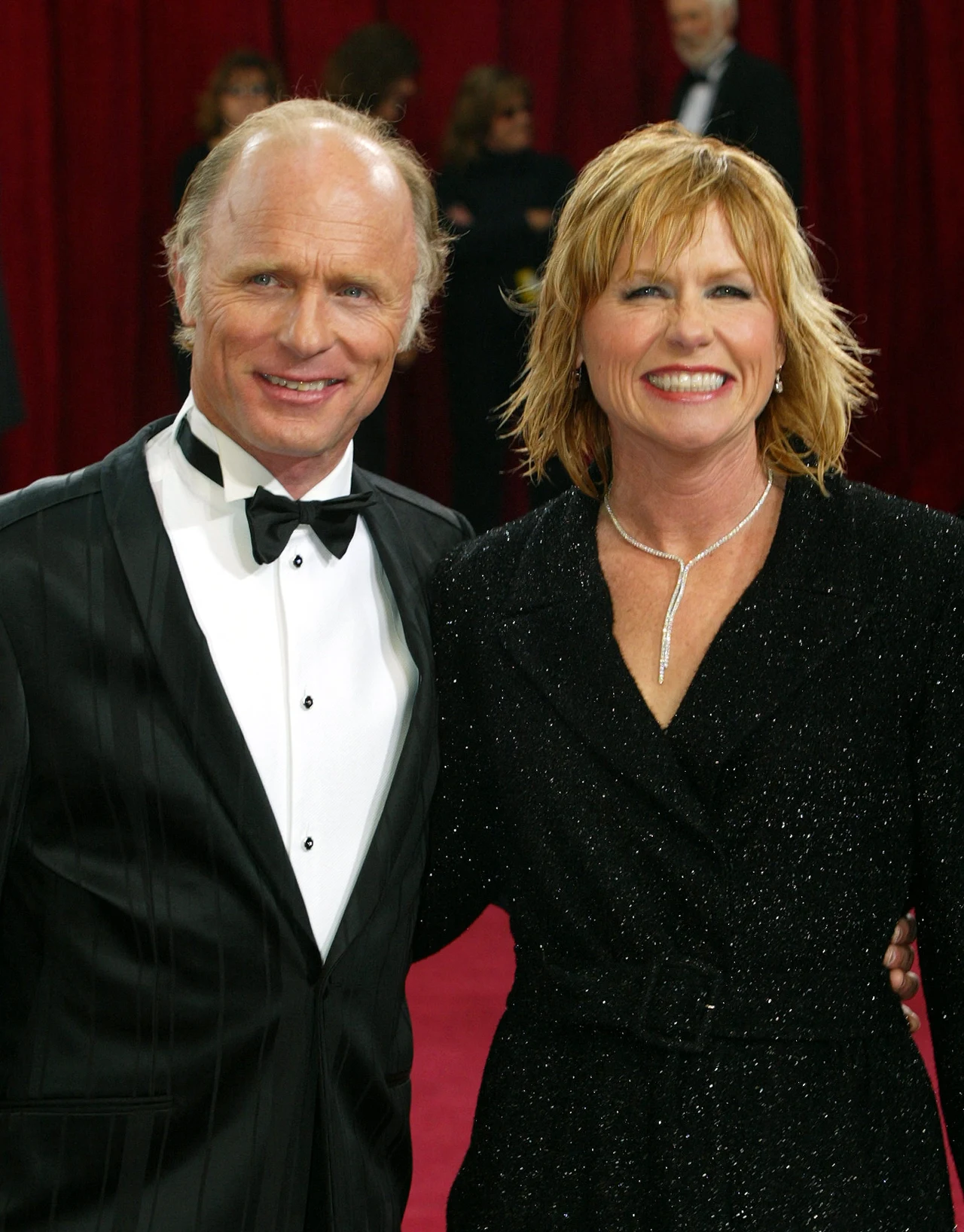 Ed Harris i Amy Madigan, 2003 r. Uśmiechnięty mężczyzna w smokingu i kobieta w czarnej sukni stoją razem na tle czerwonych kotar, pozując do zdjęcia podczas oficjalnej gali.
