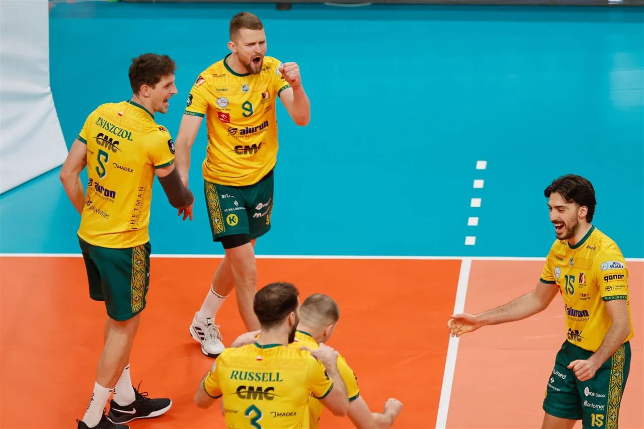 Grupa siatkarzy ubrana w żółto-zielone stroje świętuje zdobycie punktu na boisku, okazując emocje i radość; wszyscy skoncentrowani są na wydarzeniach na boisku.
