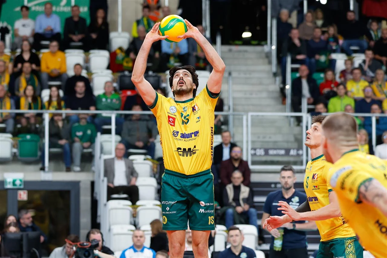 Siatkarz w żółto-zielonym stroju unosi piłkę ponad głową do wystawienia, otoczony przez innych zawodników swojej drużyny na tle kibiców na trybunach podczas meczu siatkówki.