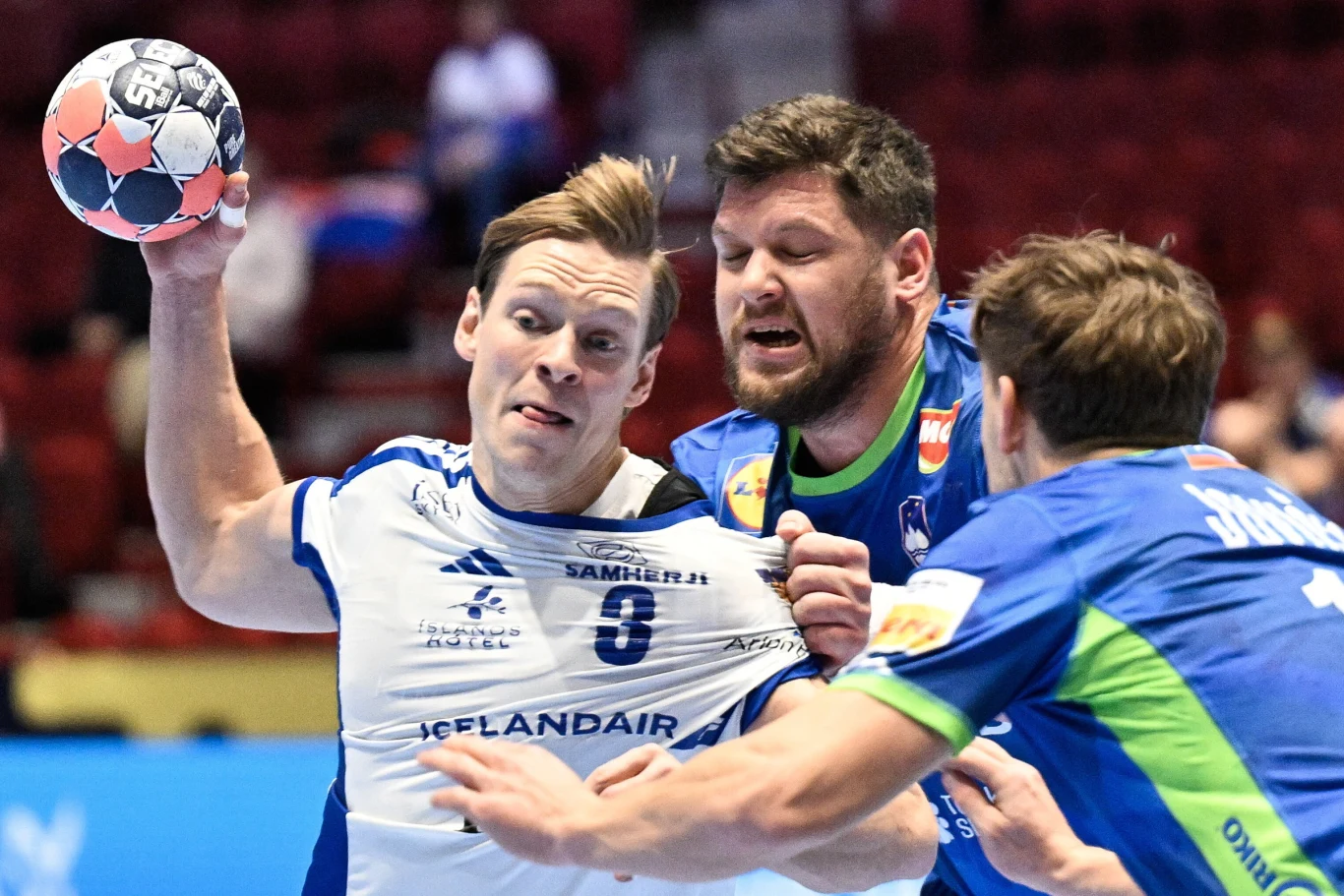 Intensywna walka trzech szczypiornistów podczas meczu piłki ręcznej, jeden z zawodników w białej koszulce trzyma piłkę i jest blokowany przez dwóch przeciwników ubranych w niebieskie stroje z zielonymi elementami.