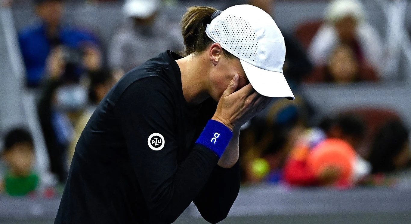 Tenisistka w czarnej koszulce i białej czapce zakrywa twarz dłońmi, wyrażając silne emocje na tle rozmazanej publiczności na trybunach.