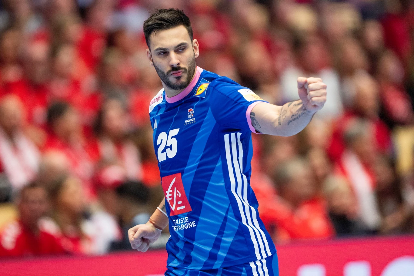 Zawodnik drużyny piłki ręcznej ubrany w niebieski strój z numerem 25 na klatce piersiowej, wykonuje gest triumfu na tle rozmytej, liczbnej publiczności w czerwonych barwach.