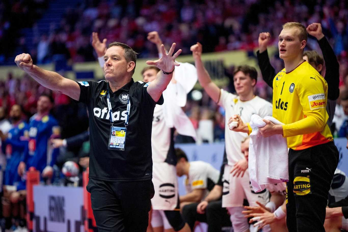 Trener drużyny piłki ręcznej wykonuje intensywny gest kierując się do zawodników lub sędziów, obok stoją zawodnicy w jednolitych strojach, w tym bramkarz w żółtym stroju, za nimi na ławce rezerwowych pozostali członkowie zespołu śledzą przebieg meczu i...