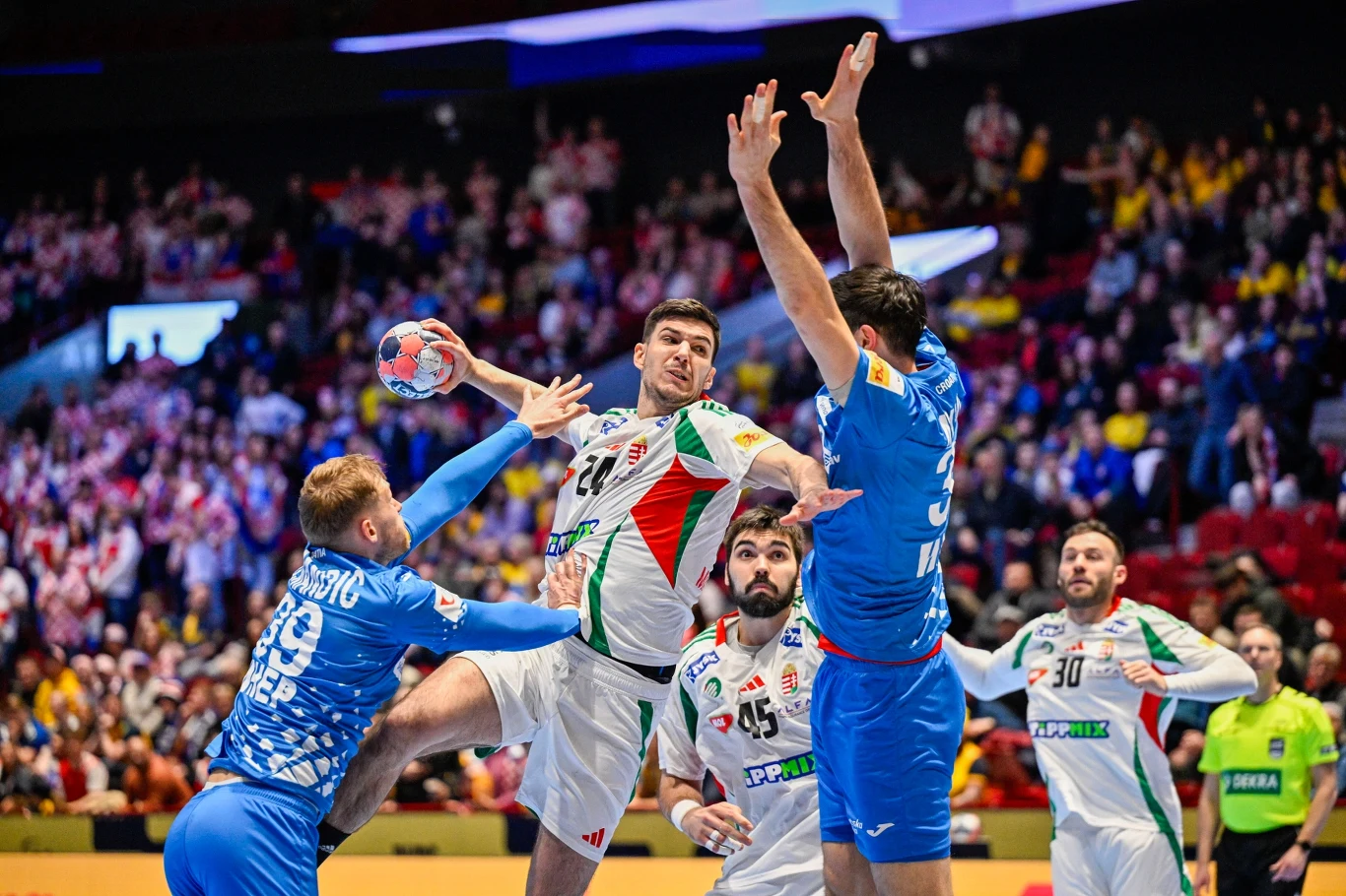 Sytuacja podczas meczu piłki ręcznej, zawodnik w białym stroju z piłką próbuje oddać rzut na bramkę, obok niego kilku graczy drużyny przeciwnej w niebieskich strojach stara się go zablokować, na drugim planie widoczny tłum kibiców i sędzia obserwujący ...