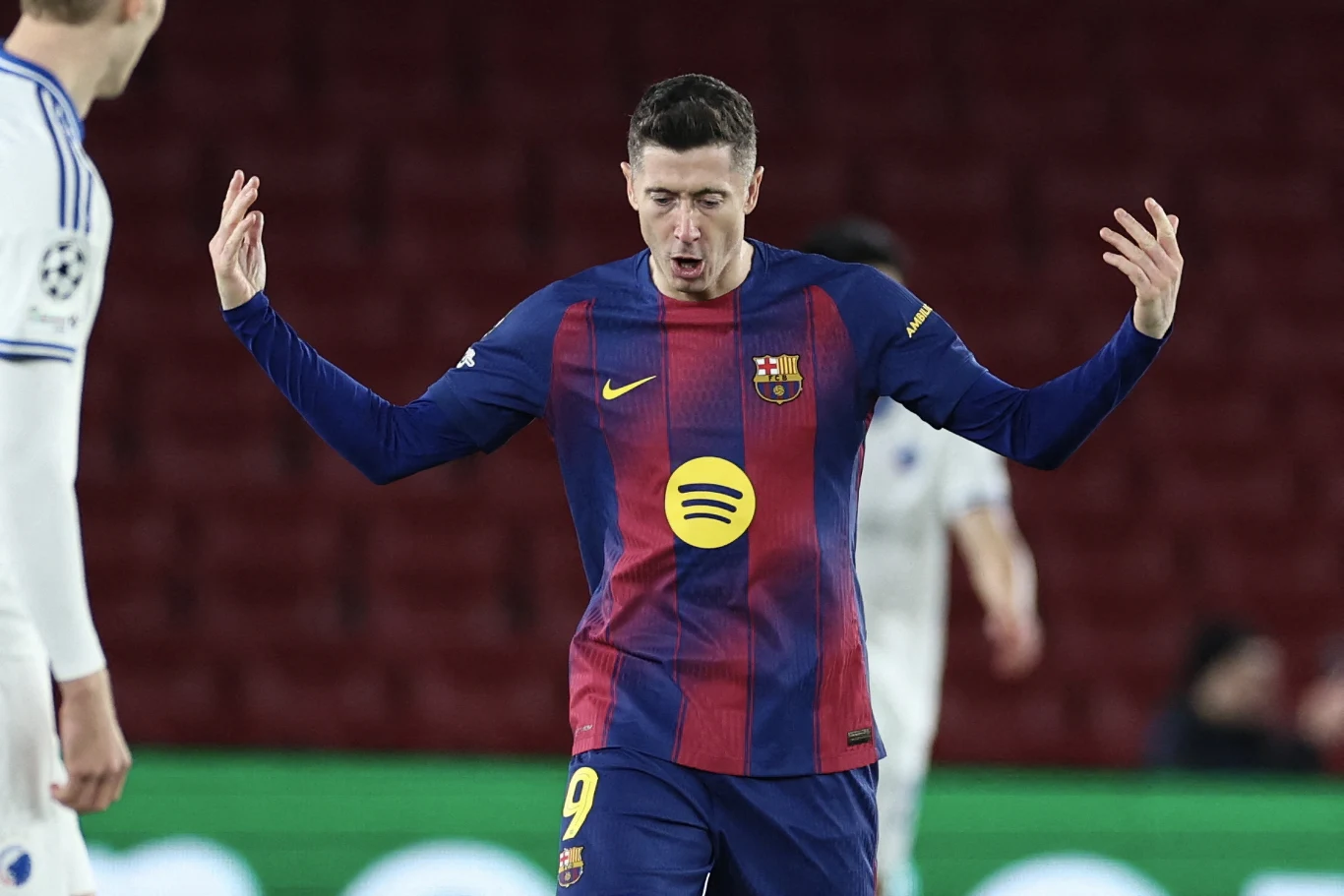 Piłkarz FC Barcelony w oficjalnym stroju meczowym z charakterystycznym logotypem na piersi unosi ramiona do góry, na pierwszym planie, na tle stadionu; w pobliżu widoczny rywal w białym stroju.