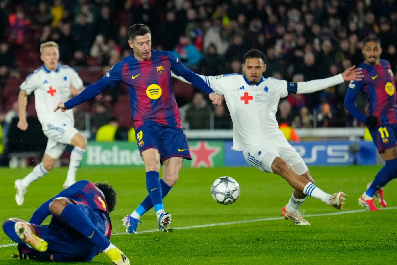 Piłkarz w bordowo-granatowym stroju próbuje zdobyć piłkę przed bramkarzem i obrońcą drużyny przeciwnej w białych strojach podczas intensywnej akcji meczowej na stadionie.