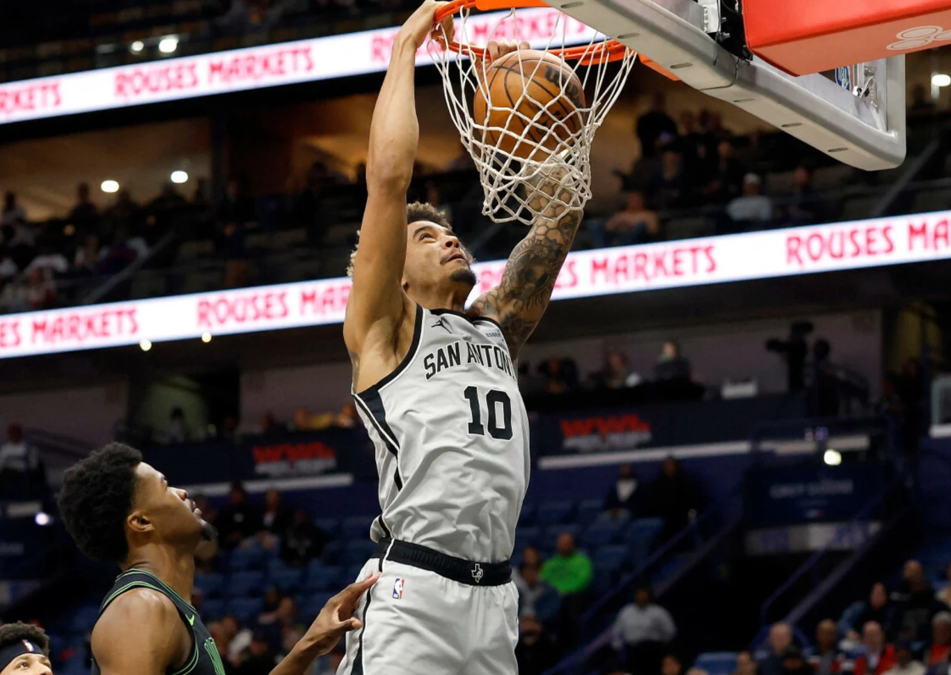 Koszykarz drużyny San Antonio Spurs wykonuje wsad do kosza podczas meczu koszykówki, a w tle widoczny jest rywal oraz trybuny z publicznością.