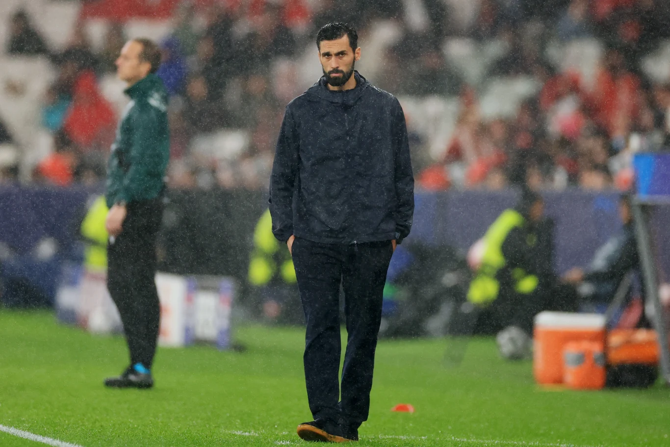 Mężczyzna w ciemnym ubraniu stoi na murawie boiska piłkarskiego, w tle widoczni są inni ludzie, kibice oraz elementy techniczne stadionu.