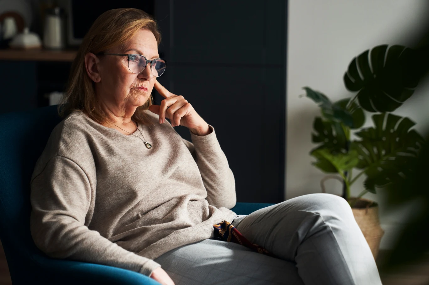 Starsza kobieta w okularach siedząca w fotelu, z ręką opartą na policzku, patrząca zamyślonym wzrokiem w bok. W tle widoczna roślina doniczkowa i ciemne meble.