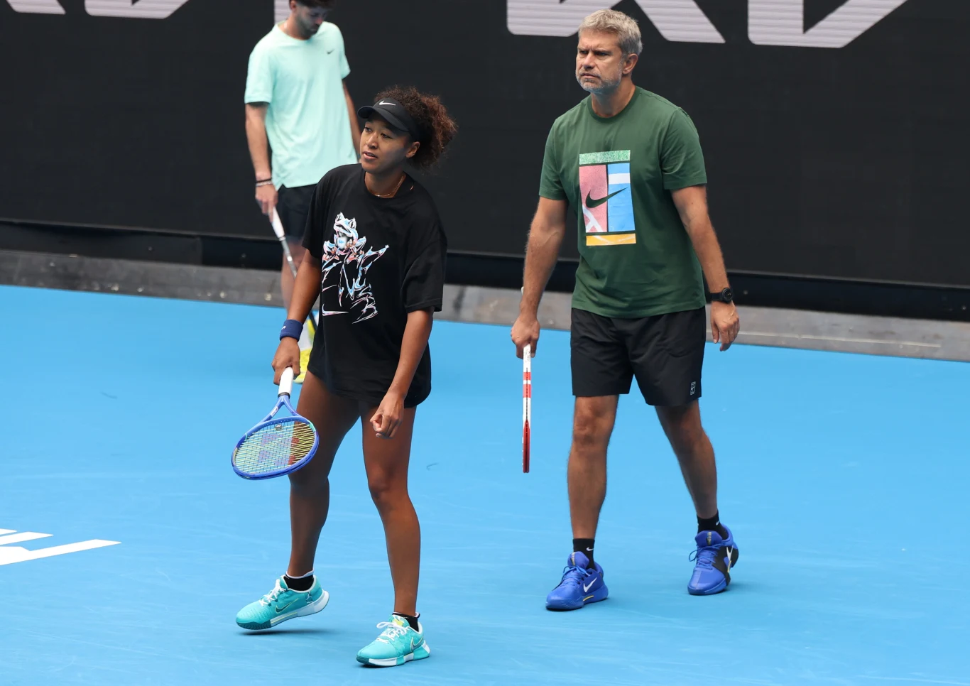 Kobieta w stroju sportowym trzymająca rakietę tenisową podczas treningu na korcie, obok niej mężczyzna również z rakietą, w tle inna osoba; dominują niebieskie kolory kortu.