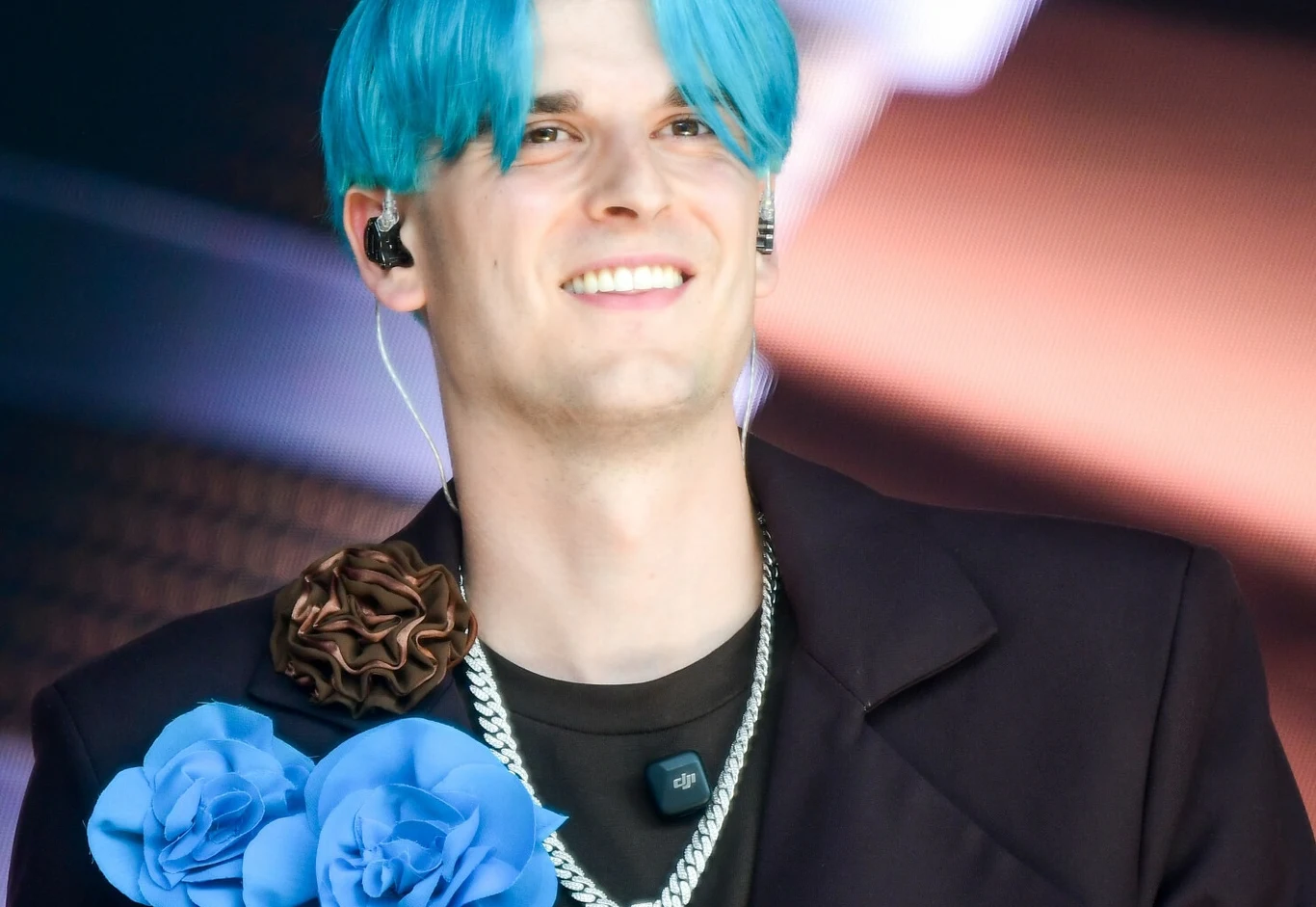 Tribbs przeprowadził się do Londynu Młody mężczyzna z niebieskimi włosami uśmiecha się, ubrany w czarną marynarkę ozdobioną niebieskimi i brązowymi kwiatami z materiału, na szyi srebrny łańcuch oraz mikrofon.