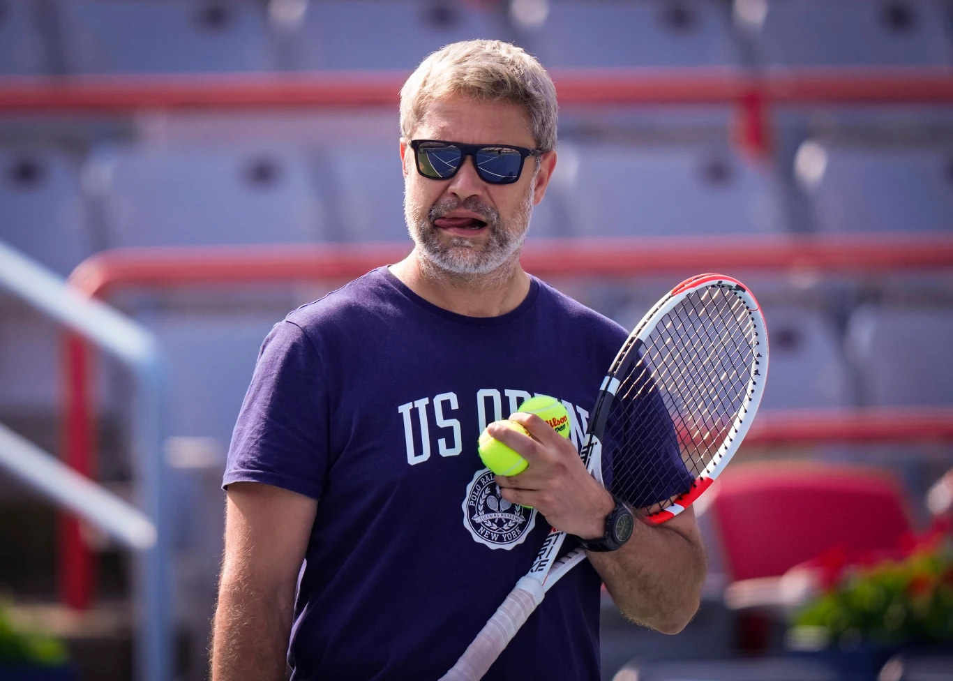 Mężczyzna w okularach przeciwsłonecznych trzymający rakietę tenisową oraz piłkę, ubrany w granatową koszulkę z napisem US Open, znajduje się na tle trybun kortu tenisowego.