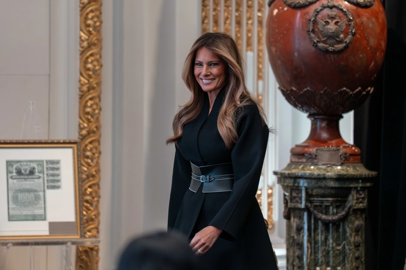 Kobieta z długimi włosami ubrana na czarno stoi w eleganckim, bogato zdobionym wnętrzu. W tle duża dekoracyjna urna na marmurowym postumencie i oprawiony dokument.