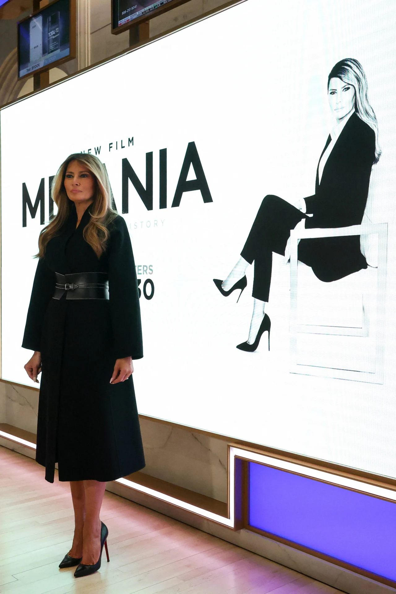 Kobieta w czarnym płaszczu i na wysokich obcasach stoi przy dużym ekranie promującym film o tytule MELANIA, na którym widnieje jej czarno-biały portret w eleganckim stroju.