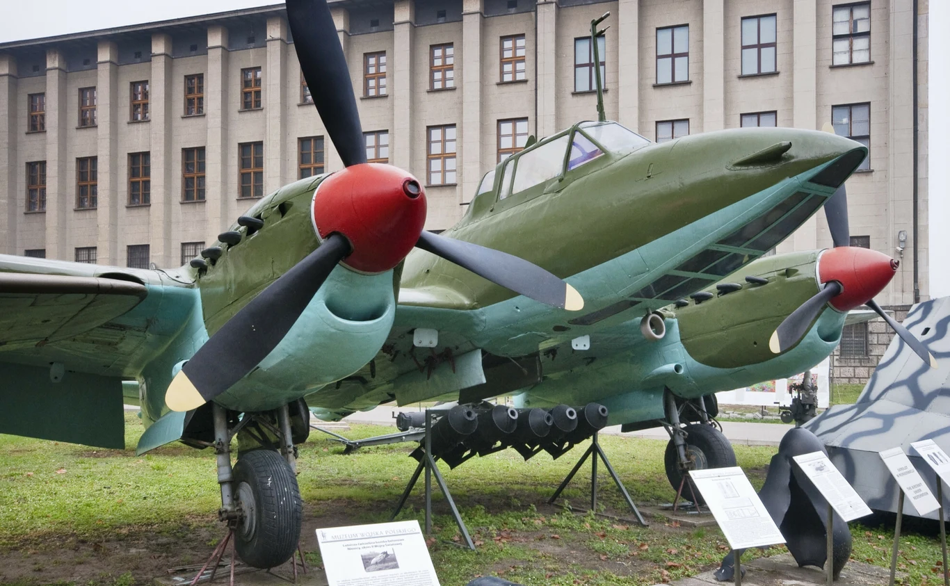 Dwusilnikowy zielono-niebieski samolot wojskowy ustawiony na zewnątrz na ekspozycji muzealnej, z widoczną armatą i bombami pod skrzydłami, na tle dużego budynku o licznych oknach.