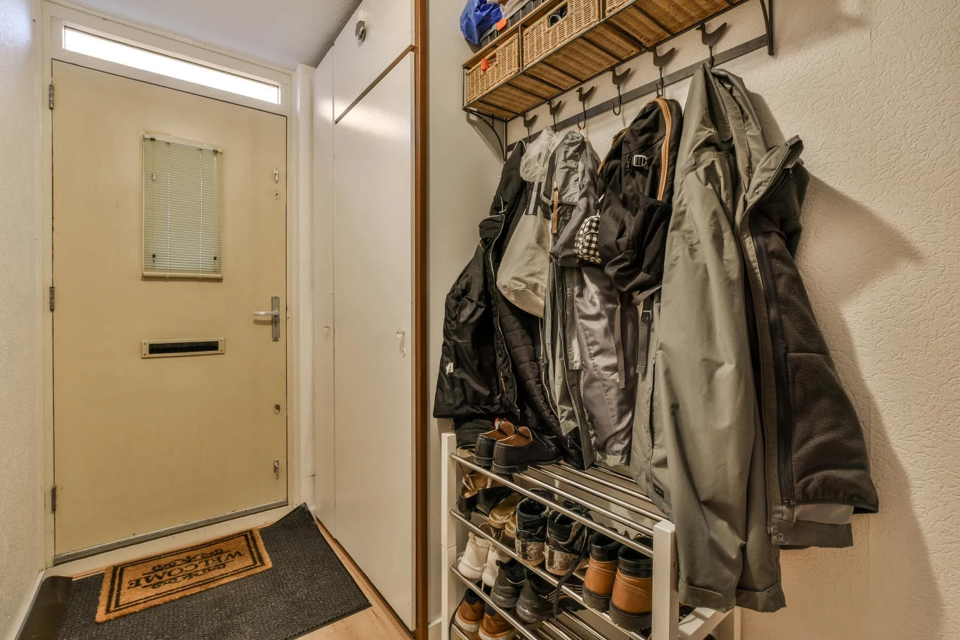 Wieszak z kilkoma kurtkami i płaszczami, poniżej metalowy stojak z ułożonymi butami, po lewej stronie zamknięte drzwi wejściowe, na podłodze leży wycieraczka.