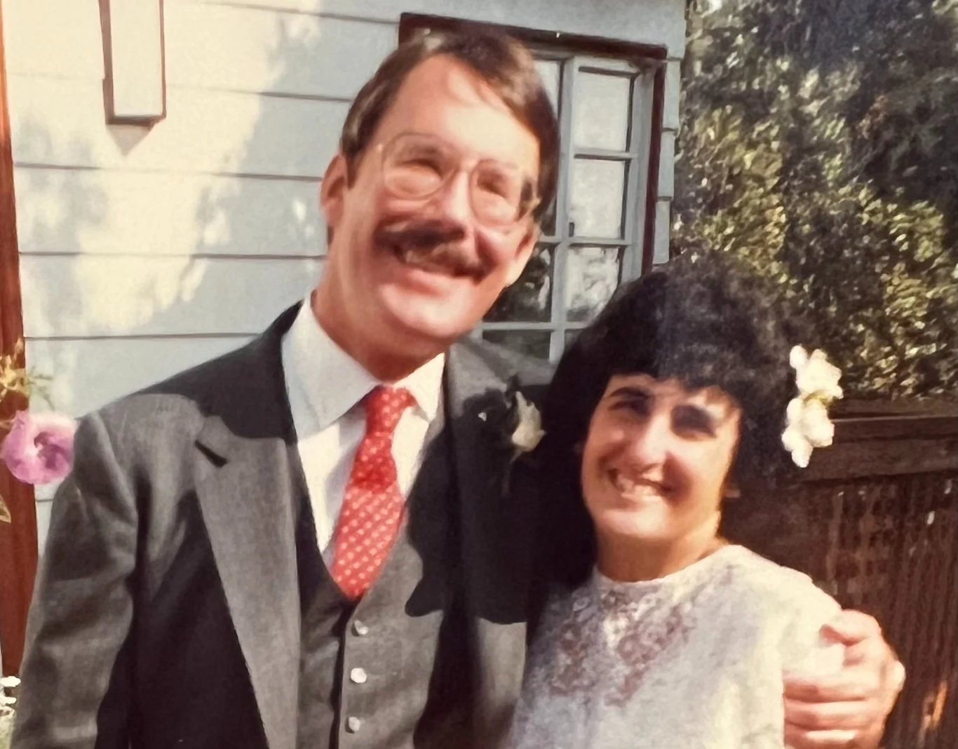 Uśmiechnięta para pozuje razem na tle drewnianego tarasu i białej ściany budynku, mężczyzna w okularach i garniturze obejmuje kobietę ubraną w białą sukienkę, ozdobioną kwiatami we włosach.