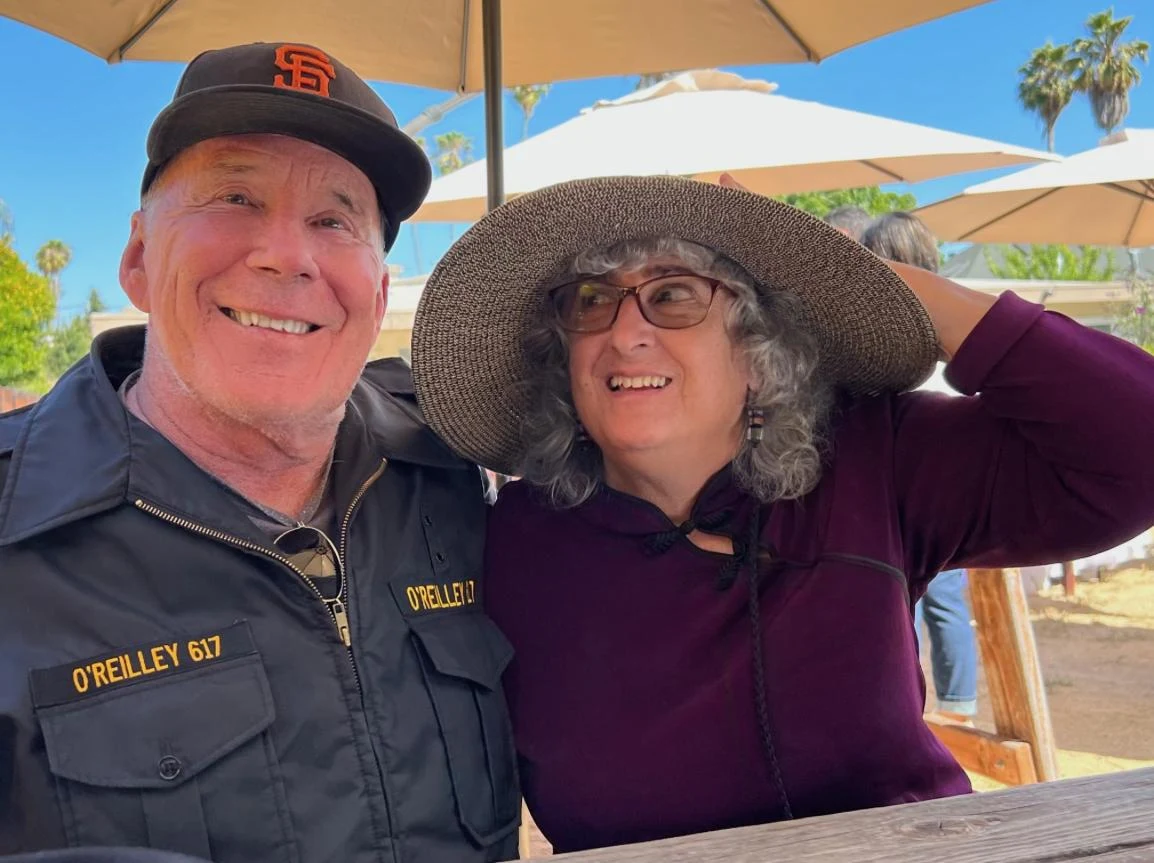 Starszy mężczyzna w czapce z daszkiem oraz kobieta w dużym kapeluszu siedzą razem przy stoliku na zewnątrz w słoneczny dzień, oboje uśmiechnięci, w tle widoczne parasole ogrodowe i roślinność.