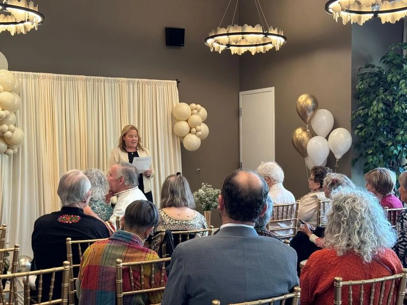 Kobieta w eleganckim stroju przemawia do grupy osób siedzących na złotych krzesłach w ozdobionej balonami i zasłoną sali, w tle widoczne są białe i złote balony oraz roślina doniczkowa, wydarzenie odbywa się w zamkniętym, nowoczesnym pomieszczeniu.