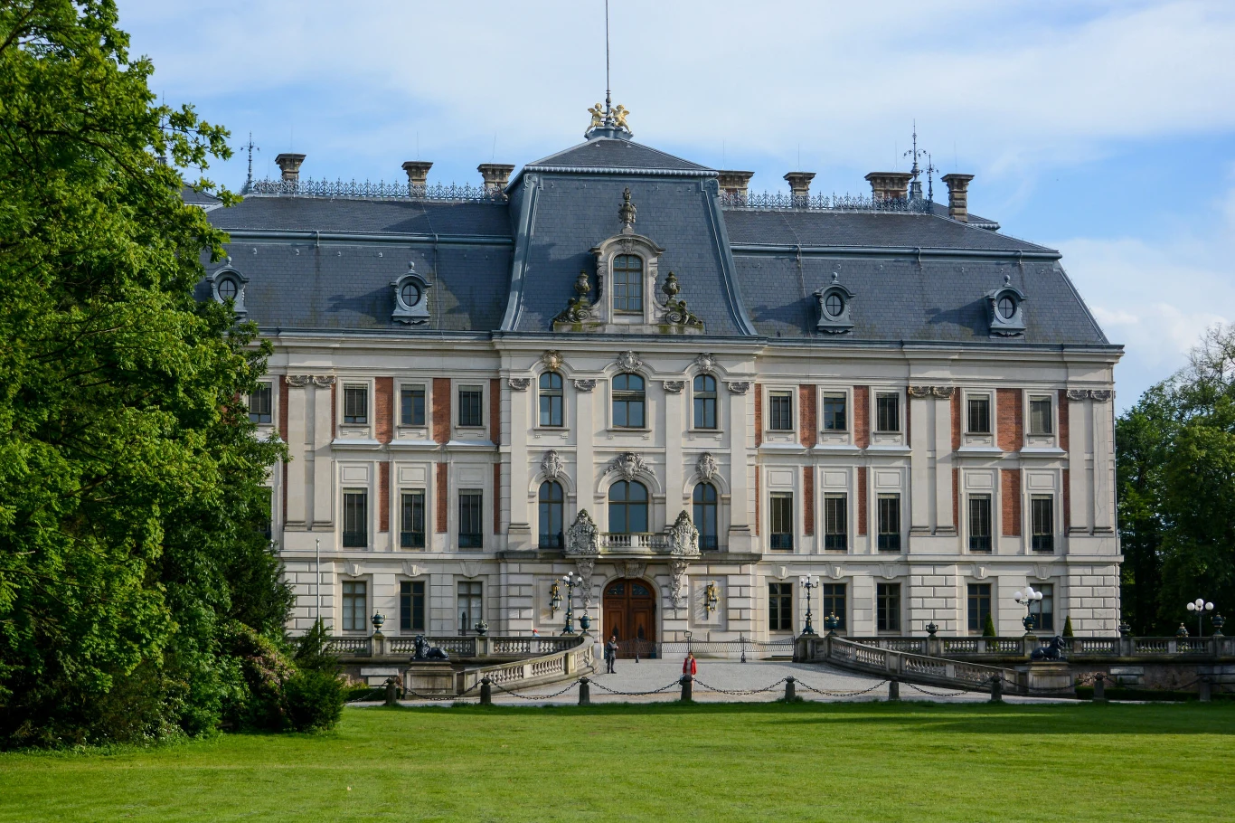Okazały pałac o bogato zdobionej elewacji, symetrycznej bryle i wysokim mansardowym dachu, otoczony wypielęgnowanym trawnikiem z drzewami po bokach.