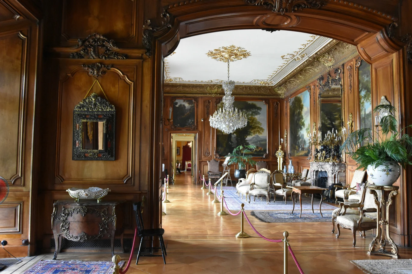 Bogato zdobione wnętrze pałacu z rzeźbionymi drewnianymi panelami, kryształowym żyrandolem i klasycznymi meblami rozmieszczonymi na perskich dywanach. Na ścianach znajdują się obrazy o tematyce pejzażowej, a całość oddzielona jest ozdobnym przejściem z...