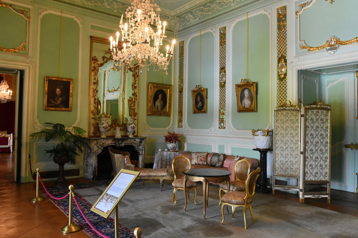 Bogato zdobiony salon z jasnymi ścianami w stylu klasycystycznym, eleganckimi meblami, kryształowym żyrandolem i portretami w pozłacanych ramach. W centralnej części salonu znajduje się stolik otoczony stylowymi krzesłami, a przy kominku widać dekoracy...