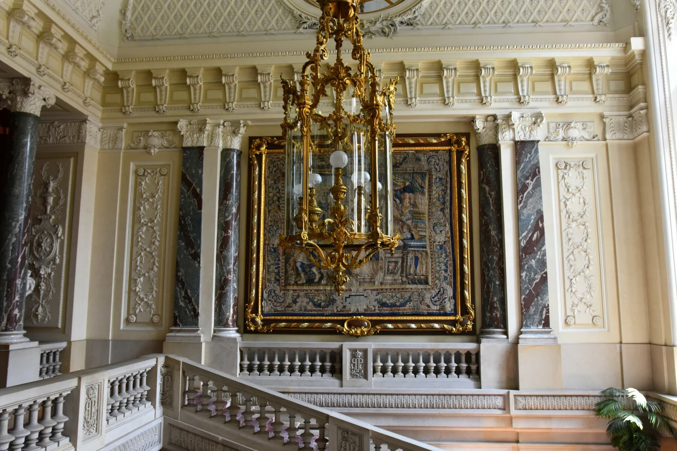 Bogato zdobione wnętrze pałacu z okazałym żyrandolem, wielobarwną tapiserią w złotej ramie oraz eleganckimi, marmurowymi kolumnami i szerokimi schodami.