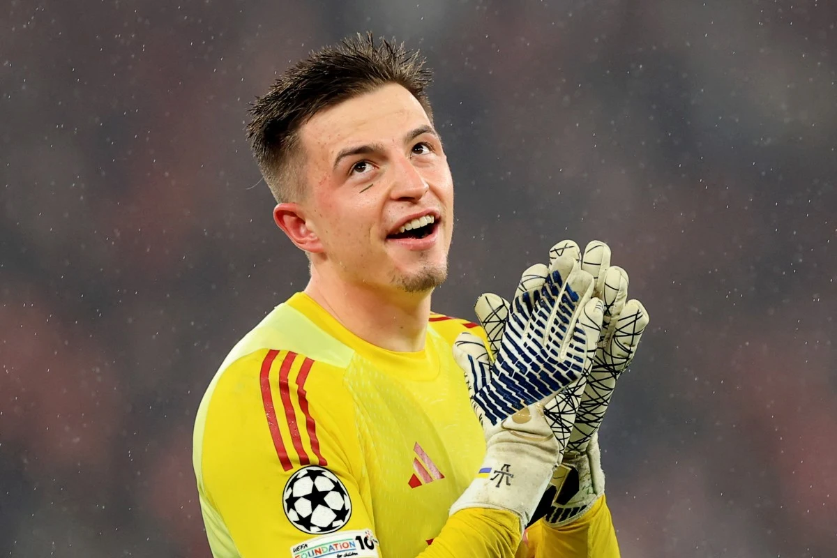 Młody bramkarz w żółtym stroju z rękawicami piłkarskimi, radosny wyraz twarzy, logo Ligi Mistrzów UEFA na rękawie, rozmyte tło stadionu.