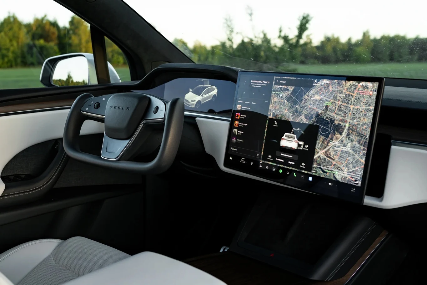 Nowoczesna kabina samochodu elektrycznego z centralnym ekranem dotykowym wyświetlającym mapę nawigacji, nietypową kierownicą o futurystycznym kształcie oraz eleganckimi, jasnymi siedzeniami. Za oknem widoczna zieleń.