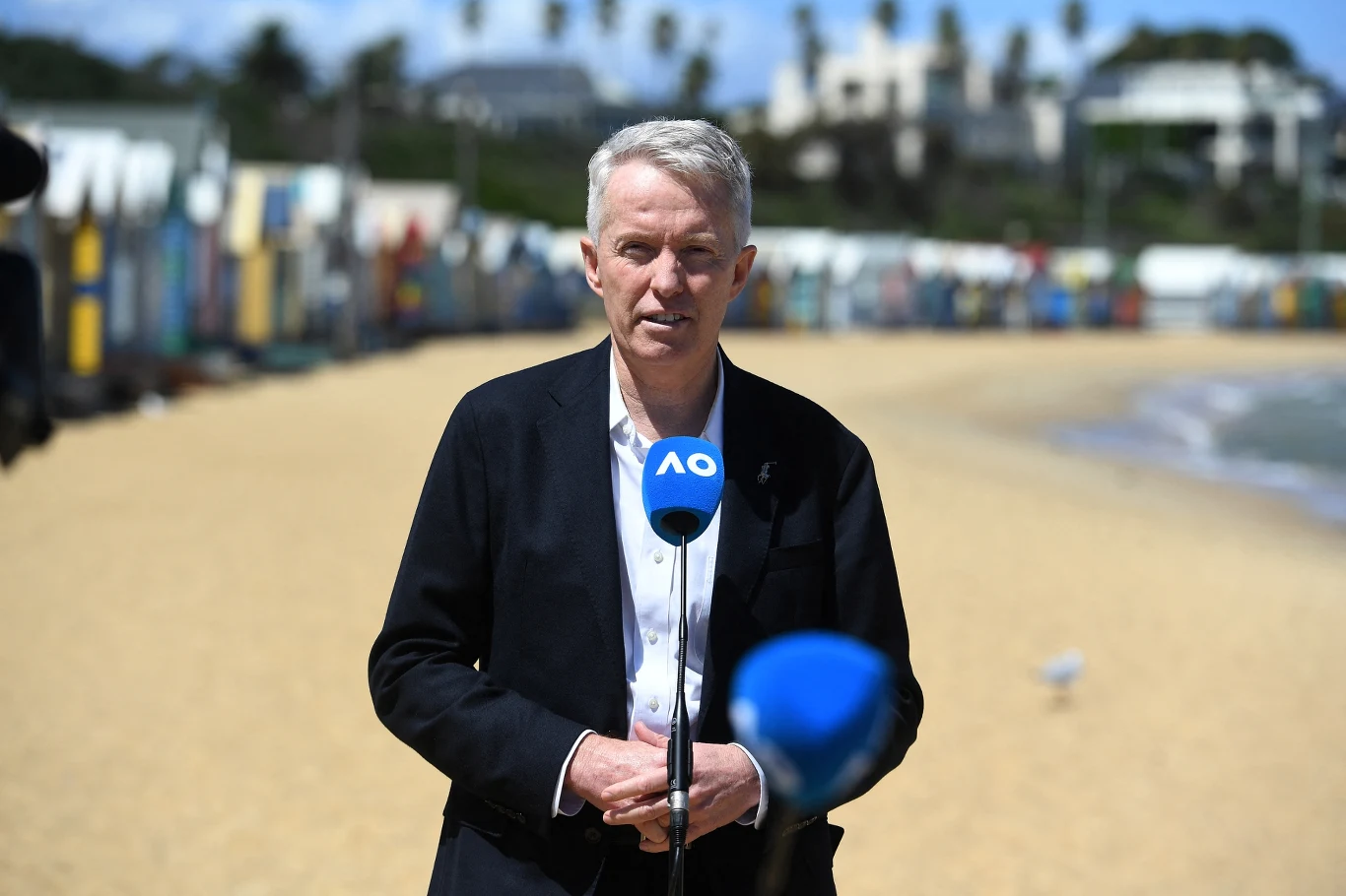Mężczyzna z mikrofonem AO udzielający wywiadu na piaszczystej plaży, w tle kolorowe domki plażowe i rozmyte budynki oraz palmy.