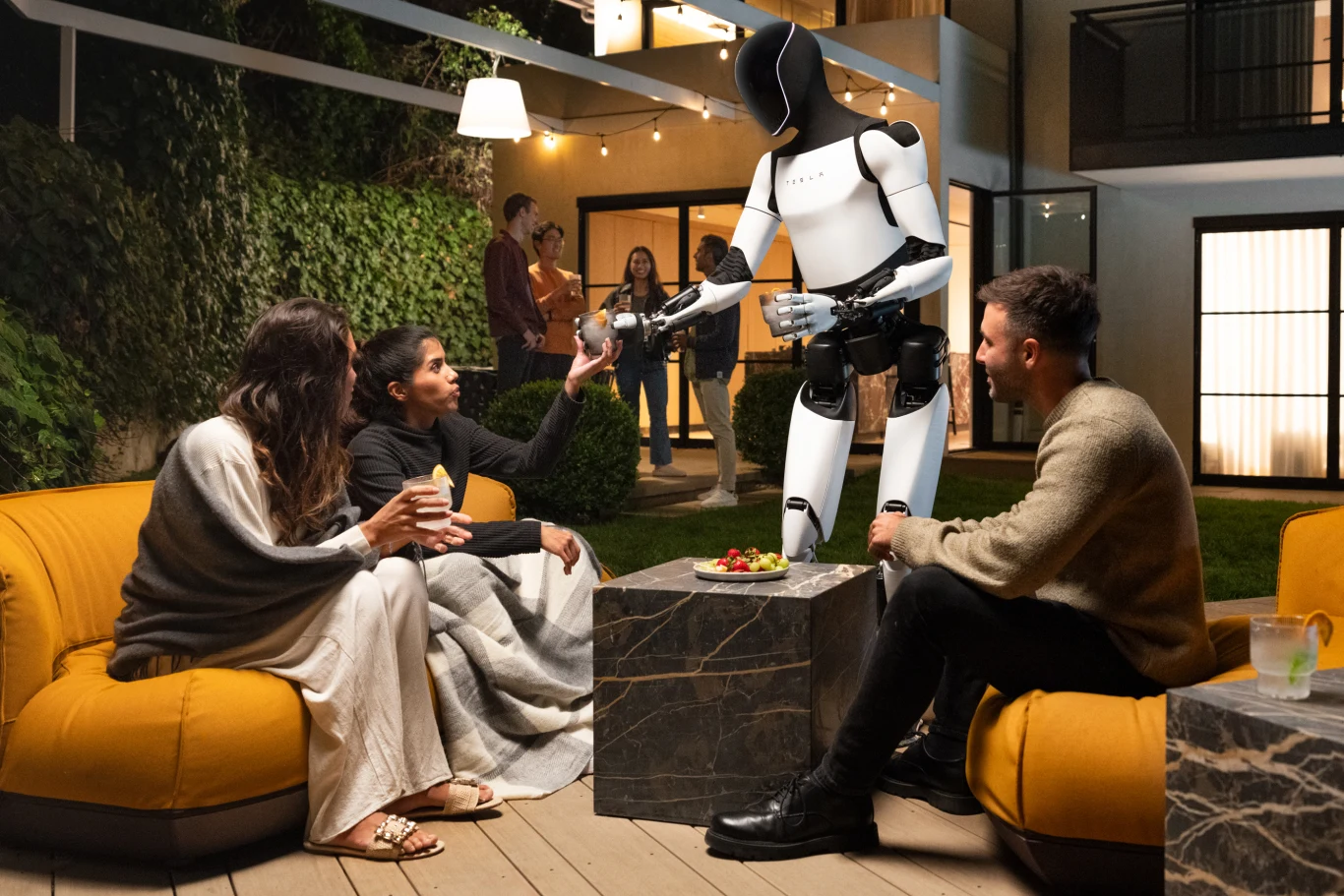 Grupa młodych ludzi siedzi na wygodnych fotelach na tarasie, a humanoidalny robot podaje napoje jednej z osób. W tle widoczne są kolejne osoby rozmawiające i przyjemne oświetlenie ogrodu.