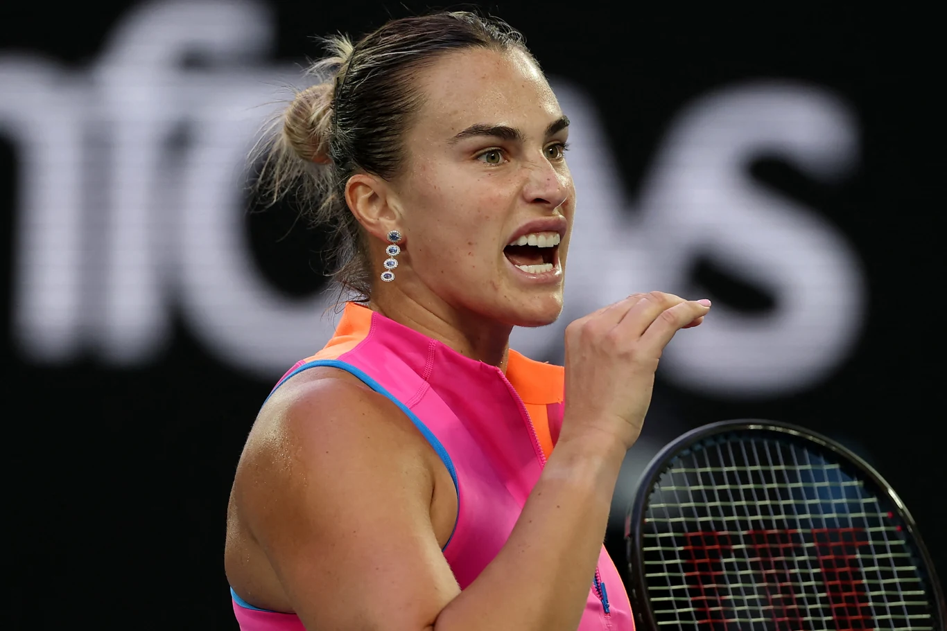 Aryna Sabalenka Tenisistka w różowo-niebieskim stroju sportowym w dynamicznym geście, z wyraźnym wyrazem emocji na twarzy, trzymająca rakietę tenisową na tle czarnego boiska.