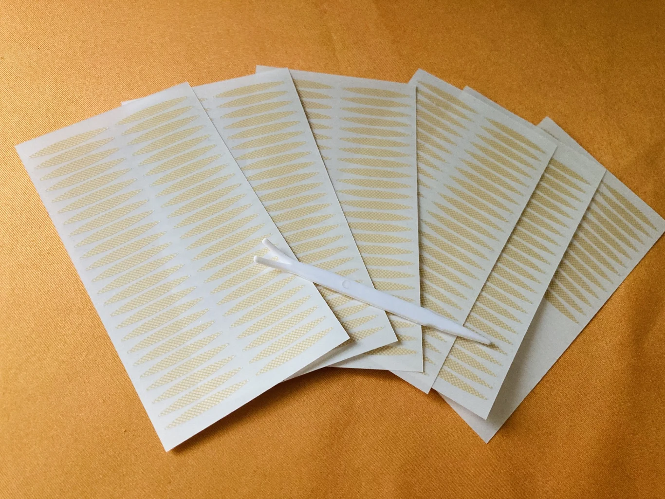 Zestaw przezroczystych, samoprzylepnych pasków do modelowania powiek ułożonych wachlarzowo na tle matowej powierzchni, wraz z białym aplikatorem umieszczonym centralnie na paskach.