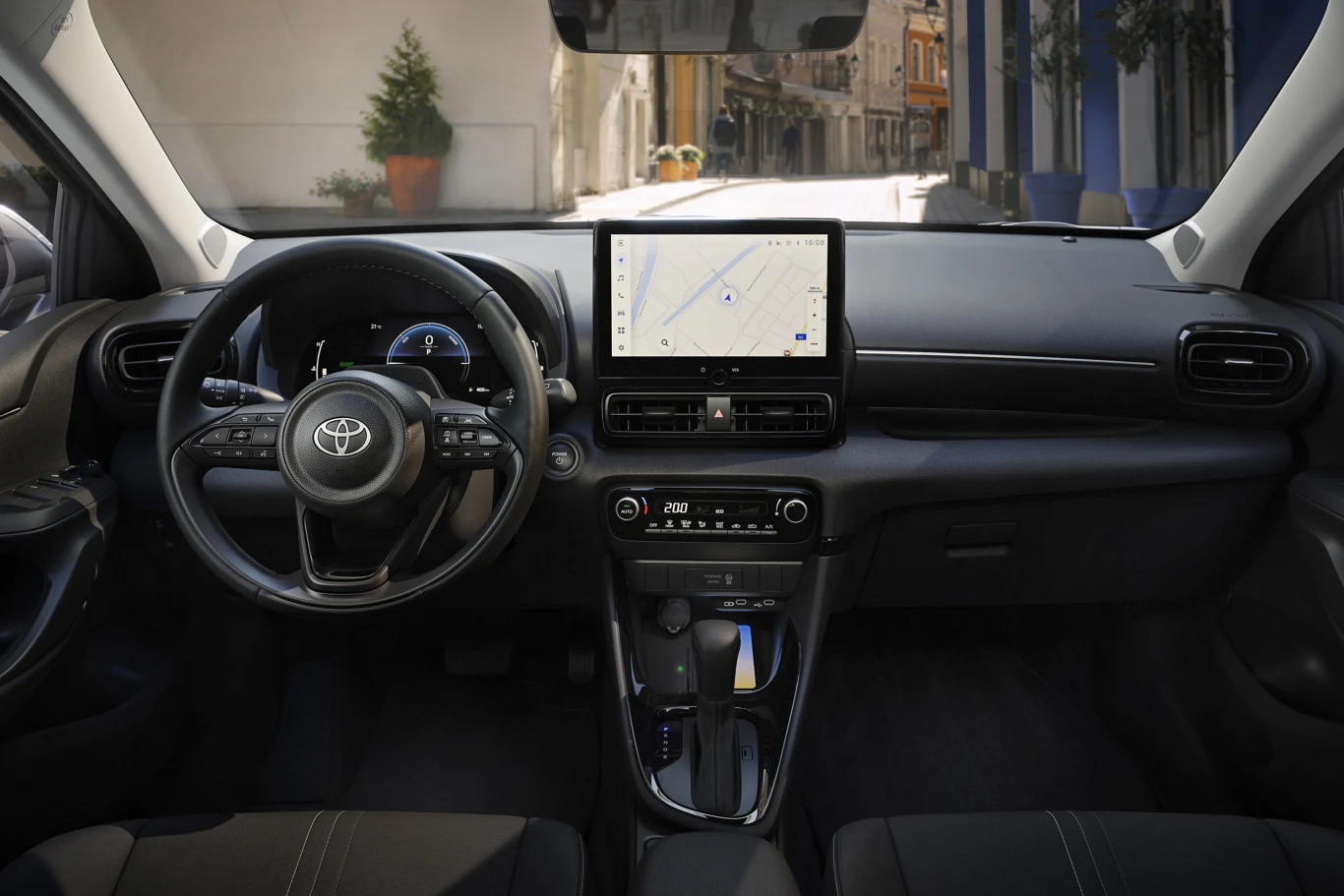 Kokpit nowoczesnego samochodu marki Toyota z dużym ekranem dotykowym na konsoli centralnej, kierownicą wielofunkcyjną, czarną deską rozdzielczą oraz panoramicznym widokiem na ulicę przez przednią szybę. Jasne, eleganckie wnętrze i wyraźnie widoczny pan...