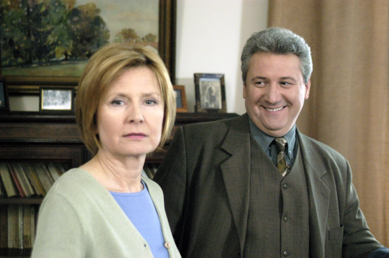 Dwie osoby w eleganckich ubraniach stoją w domowym wnętrzu, kobieta patrzy poważnie przed siebie, mężczyzna uśmiecha się szeroko, za nimi drewniany regał z książkami i zdjęciami oraz obraz na ścianie.