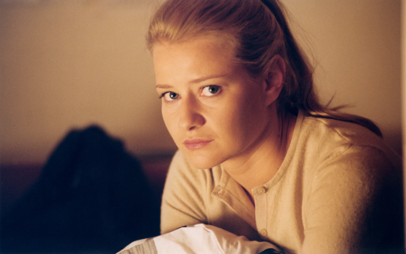 Kobieta o blond włosach w jasnym swetrze patrzy w stronę obiektywu z poważnym wyrazem twarzy, siedząc w pomieszczeniu o ciepłym oświetleniu.