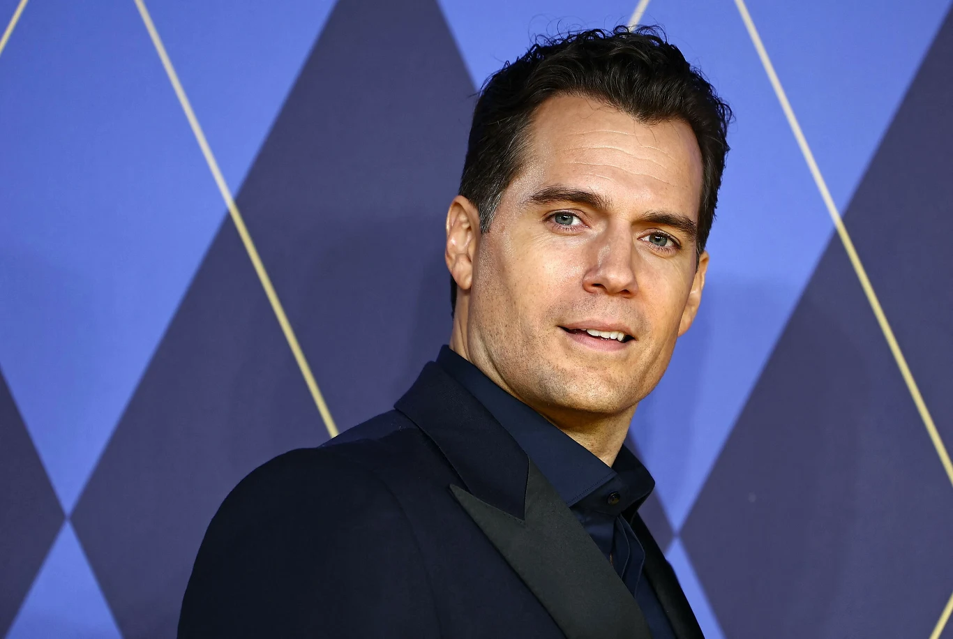 Henry Cavill Elegancko ubrany mężczyzna w ciemnym garniturze pozujący na tle niebieskich geometrycznych wzorów.