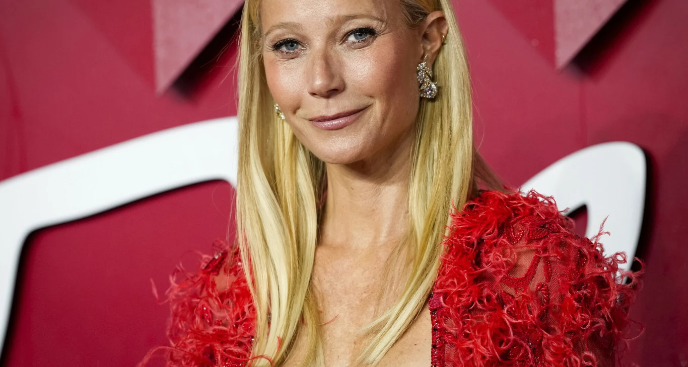 Gwyneth Paltrow - Nagrody Fashion 2023 w Londynie Kobieta z długimi blond włosami, uśmiechnięta, ubrana w efektowną czerwoną suknię z piórami, stoi na tle czerwonej ścianki z elementami graficznymi i jasnym napisem.