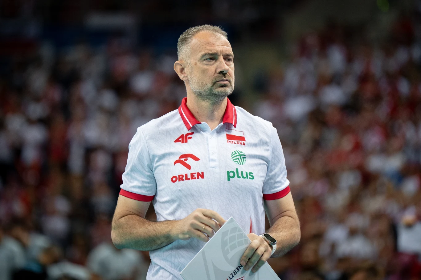 Nikola Grbić Trener siatkarski w białej koszulce reprezentacji Polski stoi na tle zamazanego tłumu kibiców na hali sportowej, trzymając w dłoniach dokumenty turniejowe.