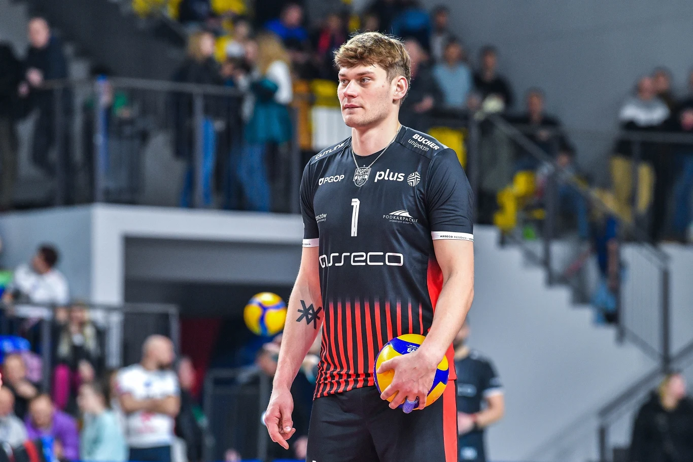 Mateusz Poręba w barwach Assseco Resovia Rzeszów Siatkarz w czarnym stroju z numerem 1 trzymając piłkę do siatkówki, stojący na boisku podczas zawodów, w tle rozmyta publiczność i fragment trybun.
