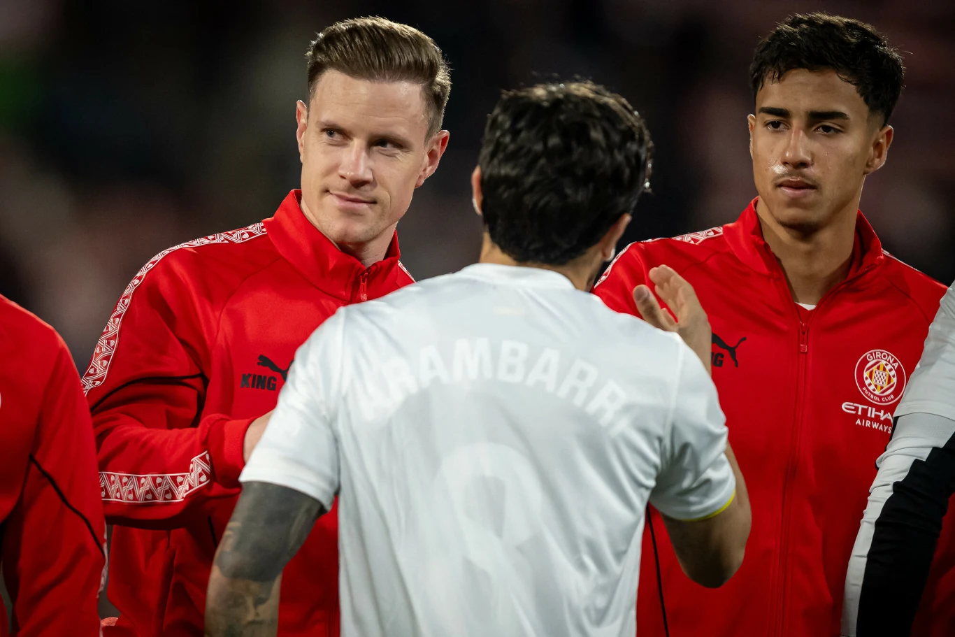 Dwóch piłkarzy w czerwonych dresach stoi blisko siebie, na pierwszym planie widoczny jest zawodnik w białej koszulce, który odwrócony jest tyłem z uniesioną ręką podczas rozmowy lub powitania.