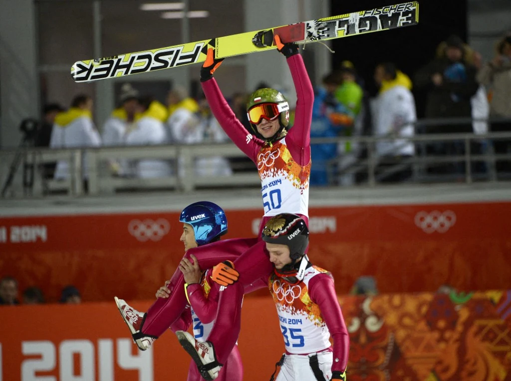 Kamil Stoch zdobył w Soczi dwa złote medale Skoczek narciarski w stroju startowym siedzi na barkach kolegów z drużyny, unosi narty w geście triumfu, wokół nich inni sportowcy i kibice na trybunach, atmosfera radości i świętowania.