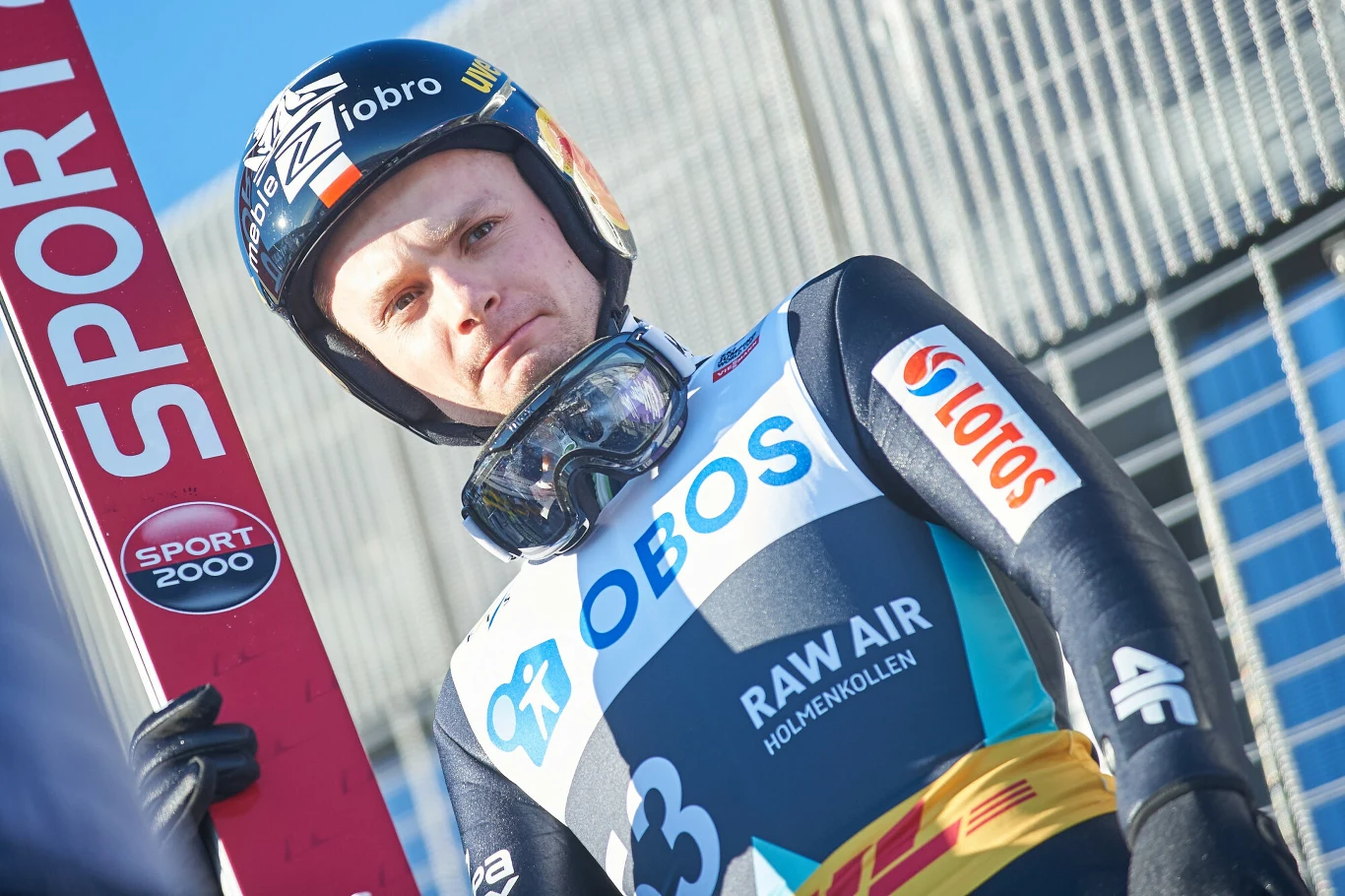 Sezon 2013/14 był najlepszym w karierze Jana Ziobro Zawodnik w kombinezonie narciarskim trzyma narty, na stroju widoczne logotypy sponsorów, na głowie kask i gogle, w tle nowoczesny budynek o przeszklonej fasadzie.