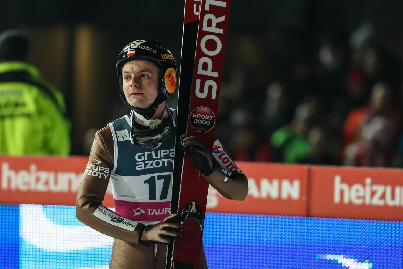Jan Ziobro zakończył karierę w 2018 roku Skoczek narciarski w kombinezonie startowym i kasku trzyma narty, stoi na tle reklam sponsorów podczas zawodów sportowych na skoczni narciarskiej.