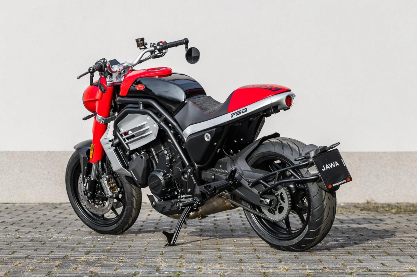 Motocykl sportowy w nowoczesnym stylu, z czerwono-czarnym lakierem, masywną tylną oponą i oznaczeniem marki na tablicy rejestracyjnej. Motocykl stoi na brukowanej nawierzchni, na tle jasnej ściany.