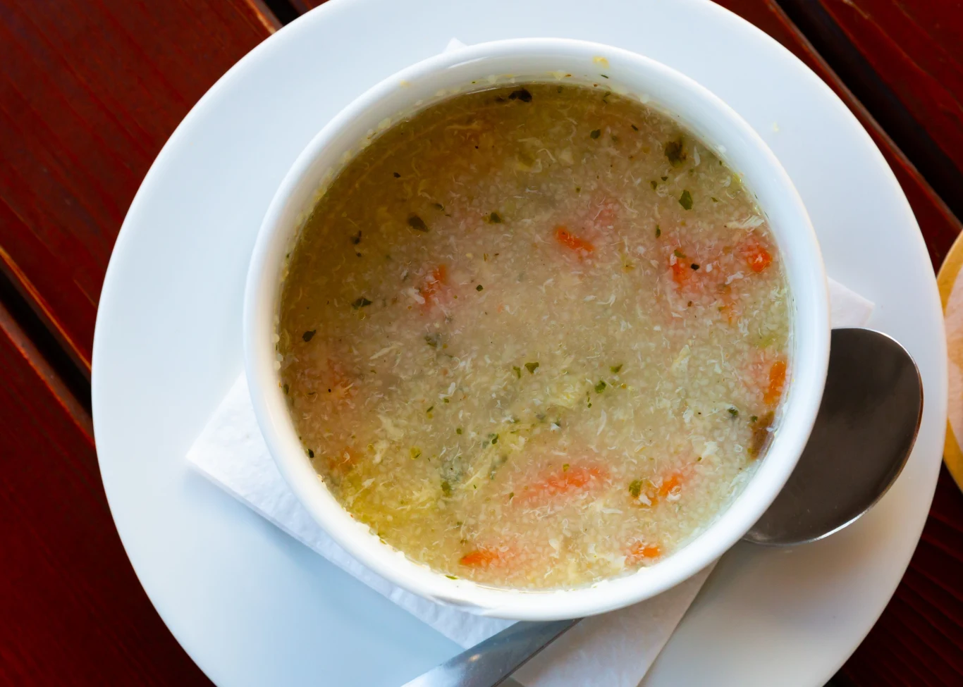 Zupa z kaszą manną jest lekkostrawna i ma łagodny smak Biała miska z gorącą zupą, w której widoczne są kawałki marchwi i zioła, stoi na białym talerzu z łyżką po prawej stronie oraz złożoną serwetką pod spodem, całość ustawiona na drewnianym stole.