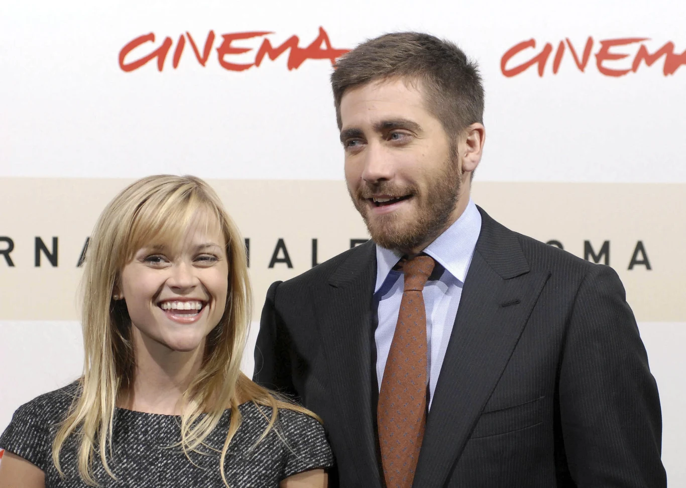 Reese Witherspoon i Jake Gyllenhaal Uśmiechnięta kobieta w jasnej sukience stoi obok mężczyzny w garniturze z krawatem, oboje znajdują się na tle napisu CINEMA, wyglądają na zadowolonych i zrelaksowanych.