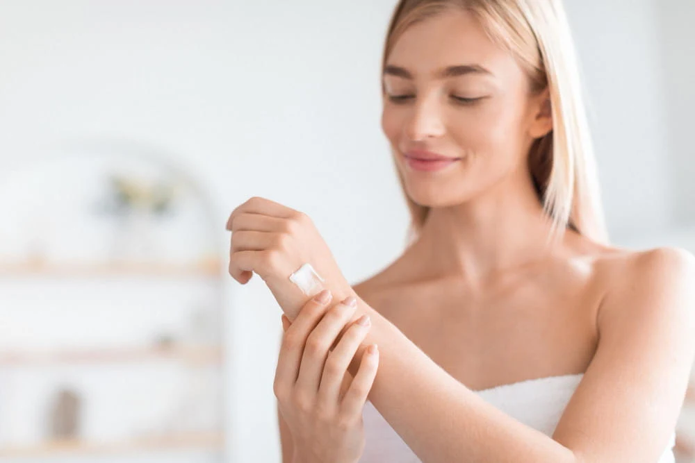 Młoda kobieta z jasną cerą nakłada krem na przedramię, delikatnie się do siebie uśmiechając, znajduje się w jasnym, domowym wnętrzu.